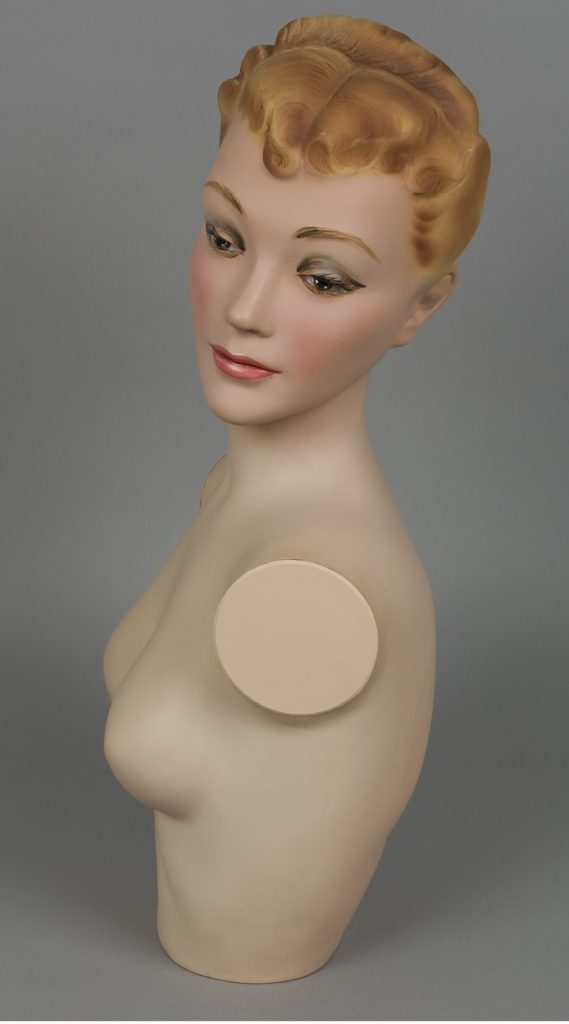 Composite shop dummy head, shoulders and bust only with head facing left side with blond hair, pink lipstick and green and brown eye shadow. There are metal plates at armholes to attach arms but no arms.